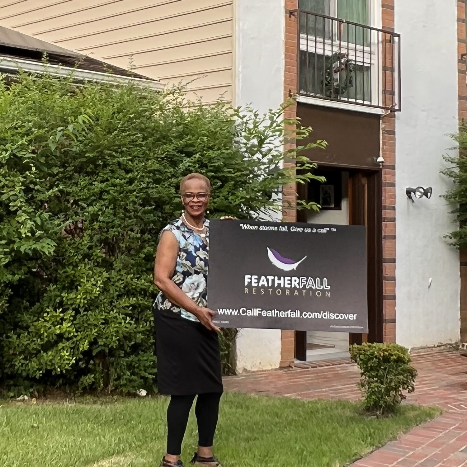 A happy customer standing in front of home holding Featherfall sign after roof repairs and siding replacement.